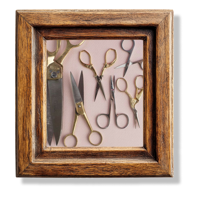 Six different shaped scissors of different sizes on a pink tablecloth in a wooden frame.